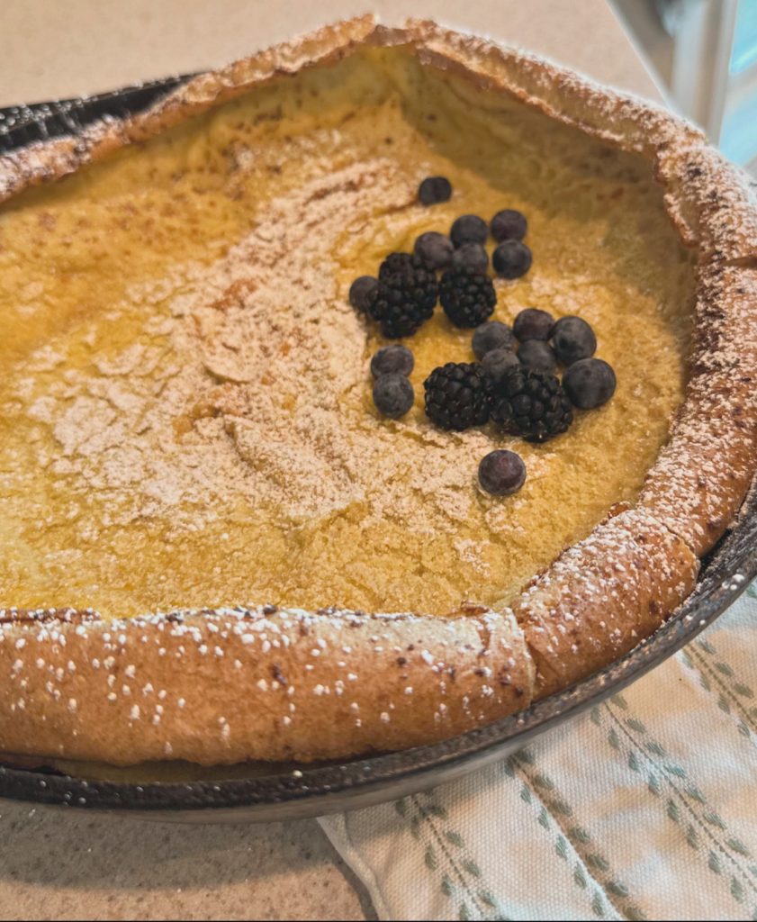 Dutch baby pancake in cast iron pan with berries and confectioners sugar on top setting on kitchen counter.