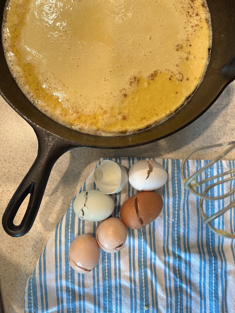 dutch baby batter in hot pan next to egg shells and hand mixer on kitchen towel. 
