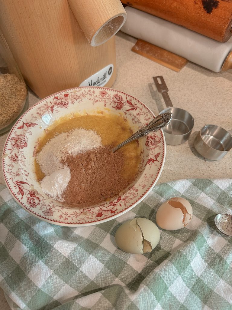 brownie ingredients in pink and white bowl ready to mix on counter beside grain mill, measuring cups and egg shells on green and white towel