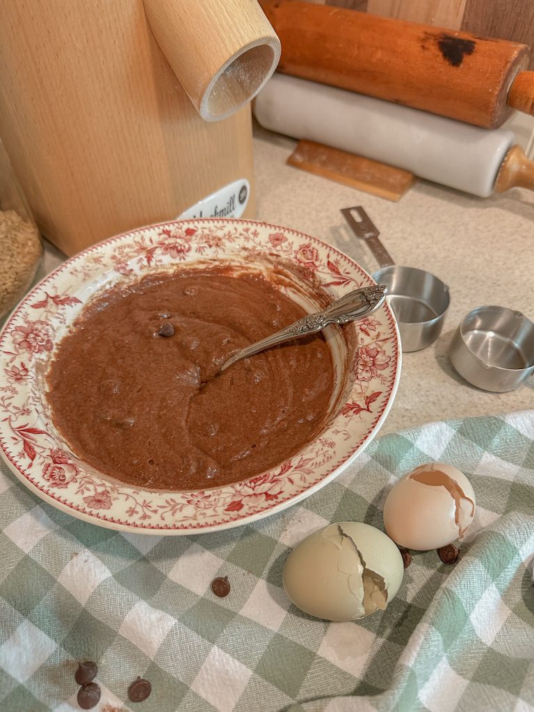 brownie batter mixed in pink and white bowl beside grain mill and egg shells on green and white towel