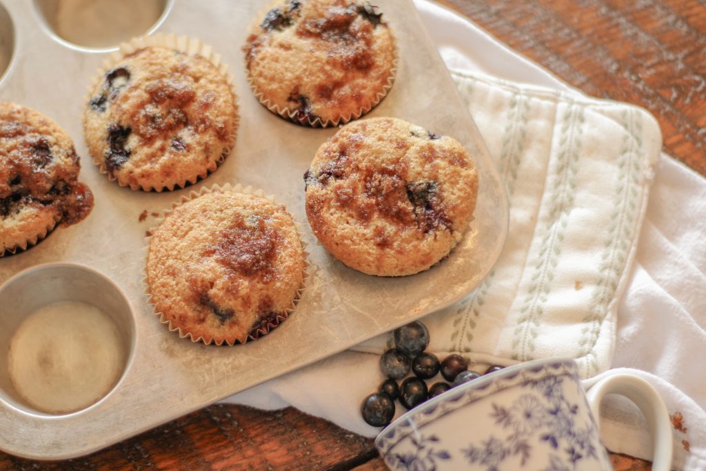 Blueberry muffins with streusel topping in silver muffin tin setting on green and white pot holder on wooden table with blue and white cup of spilled blueberries