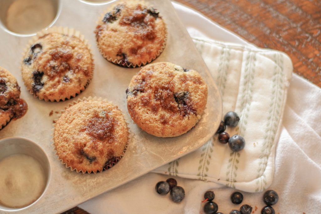 Blueberry muffins with streusel topping in silver muffin tin setting on green and white pot holder on wooden table with spilled blueberries