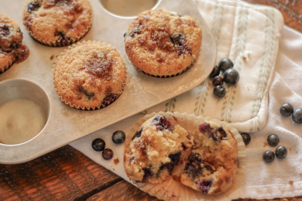 Blueberry muffins with streusel topping in silver muffin tin setting on green and white pot holder on wooden table with spilled blueberries