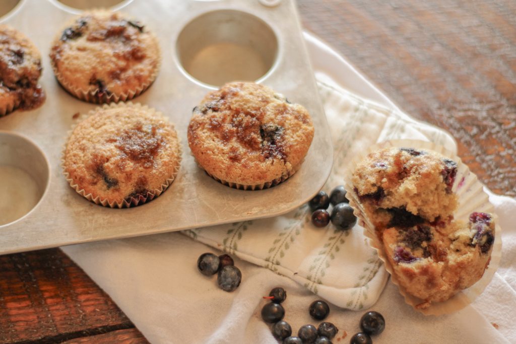 Blueberry muffins with streusel topping in silver muffin tin setting on green and white pot holder on wooden table with spilled blueberries