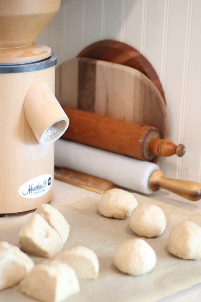 Dough for easy homemade bagels with freshly milled flour partially formed into balls setting on parchment paper on counter beside rolling pens, cutting boards, and grain mill