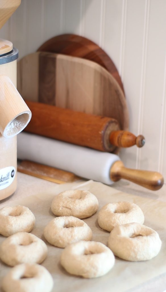 Easy homemade bagels with freshly milled flour shaped and setting on parchment paper on counter beside rolling pens, cutting boards, and grain mill