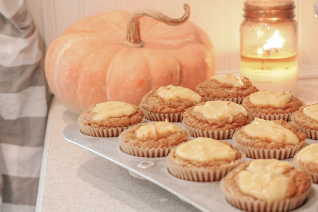 Pumpkin muffins with maple cream cheese in muffin tin setting on counter in front of orange pumpkin and lit candle