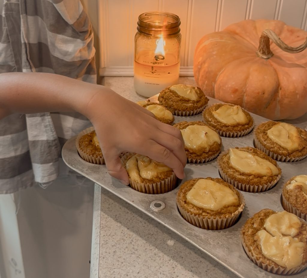 Pumpkin muffins with maple cream cheese in muffin tin setting on counter in front of orange pumpkin and lit candle with child's hand grabbing a muffin