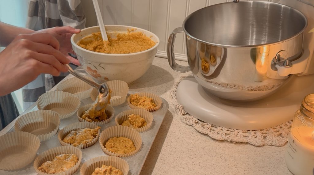 Filling muffin tin on counter top beside stand mixer and candle with pumpkin muffin batter