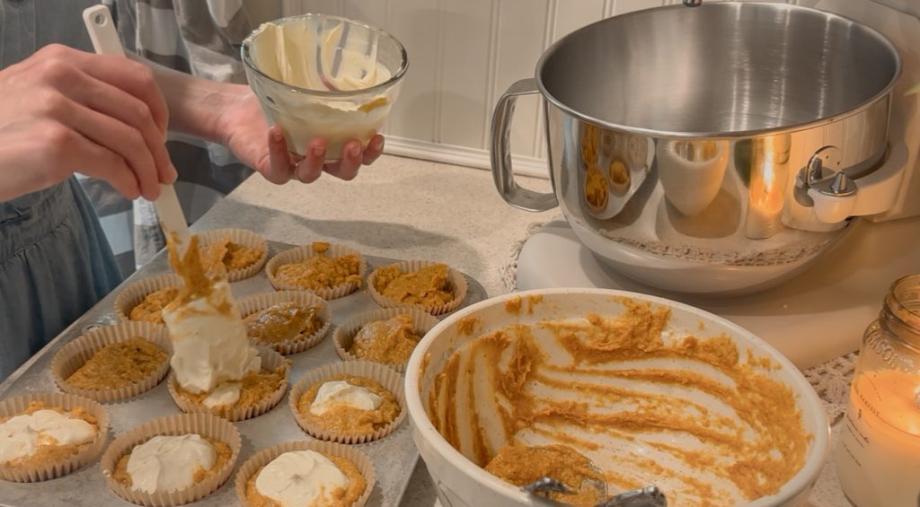 Lady adding maple cream cheese to muffins in muffin tin on counter beside mixing bowls and candle 